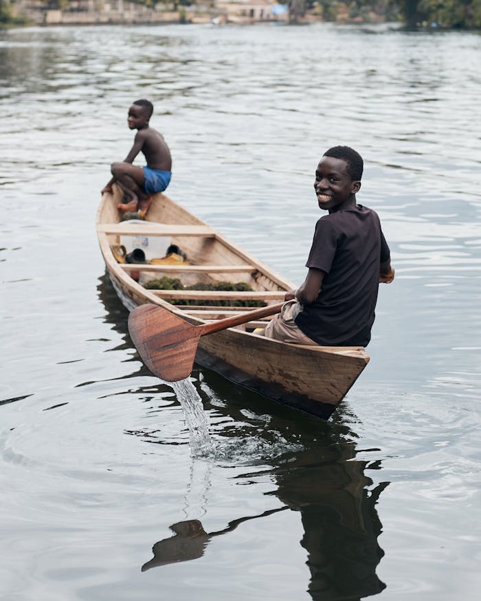 menu-07 Two boys smiling while rowing a wooden boat on a peaceful river, enjoying a day outdoors.